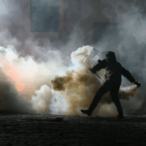 A person in riot gear with a shield walks through thick smoke on a cobblestone street at night,
