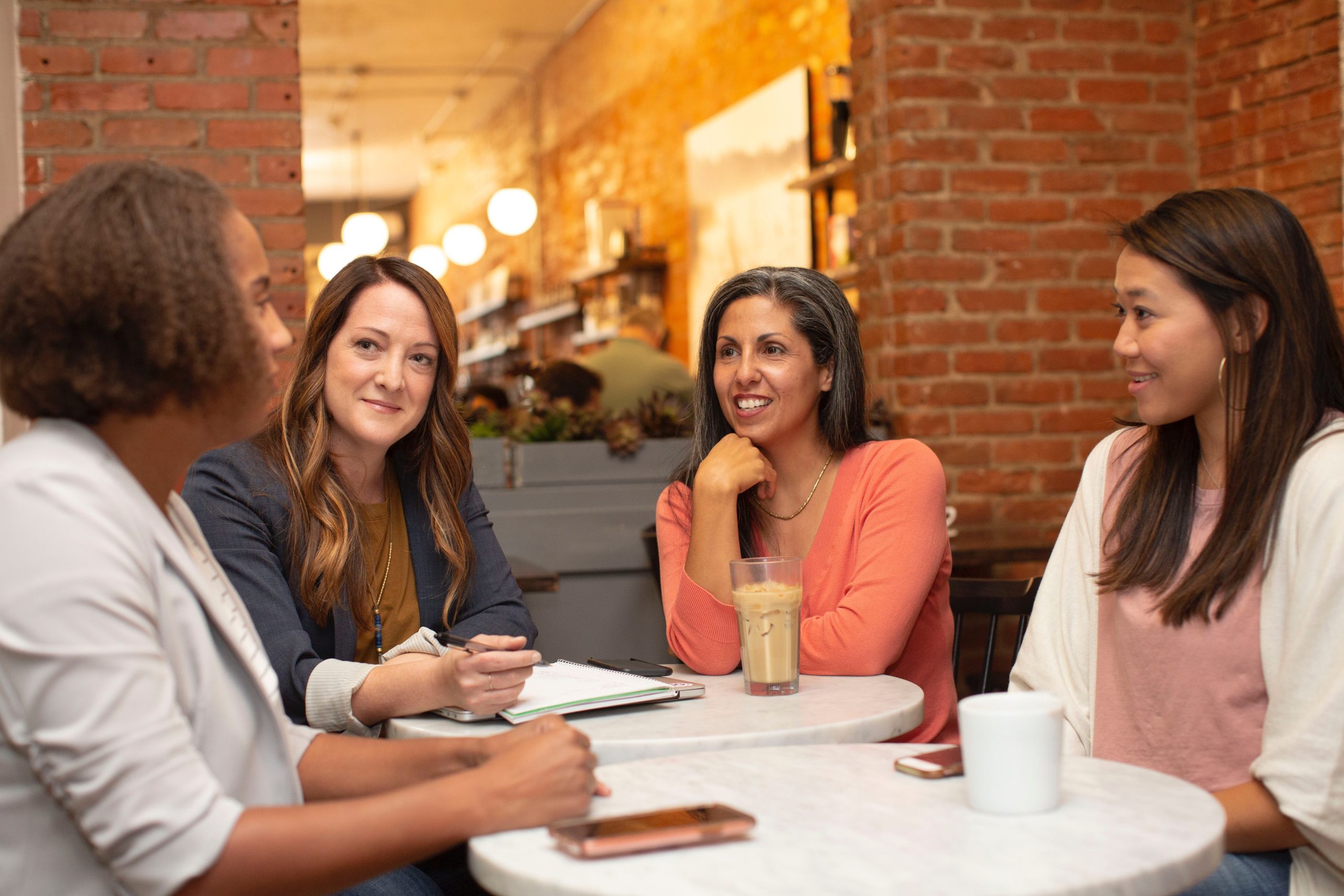 Four diverse women engaging in a relaxed conversation at a cafe, highlighting themes of DEI and wellbeing in the workplace.